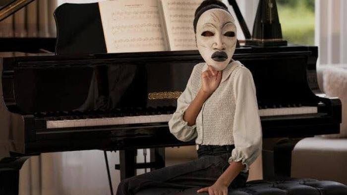 Strung: person in a white mask sitting by a piano in a well-lit room.