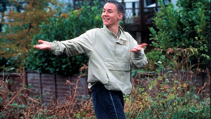 Sweet Sixteen: A young White male teen in a beige jacket smiles as he holds his arms out to the side, walking through a housing estate