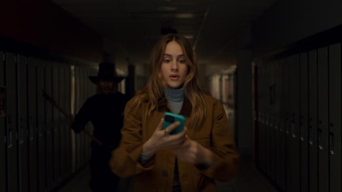 Thanksgiving Movie: Teen Jess (Nell Verlaque) stands in a dark hallway looking at her phone while a figure in a pilgrim hat, holding an axe, sneaks up behind her