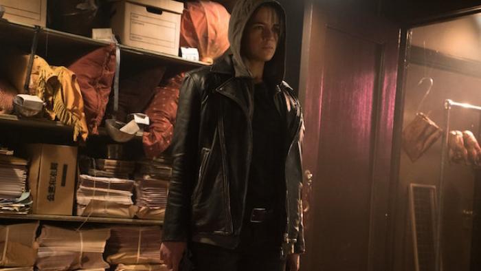 The Assignment Movie: Michelle Rodriguez stands inside a back room wearing a hoodie, storage files and rotisserie chickens in the background.