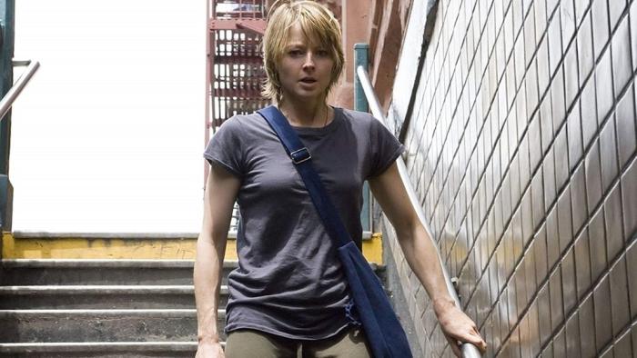 Jodie Foster walks down a subway staircase in daylight wearing a t-shirt and a blue messenger bag. 