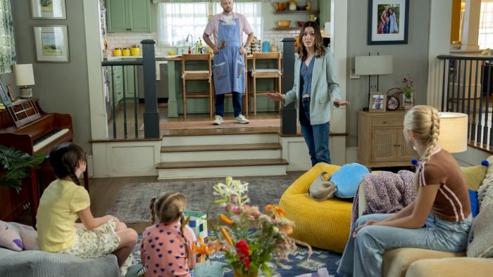 The Breadwinner movie: Mandy Moore talks to her three daughters in the living room while Nate Bargatze is in the kitchen wearing an apron