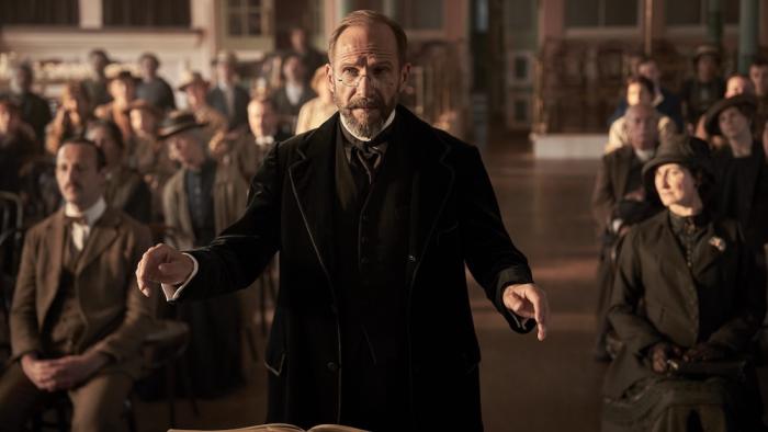 The Choral movie: A bearded Ralph Fiennes in black coat and glasses gestures while speaking to seated audience in period setting