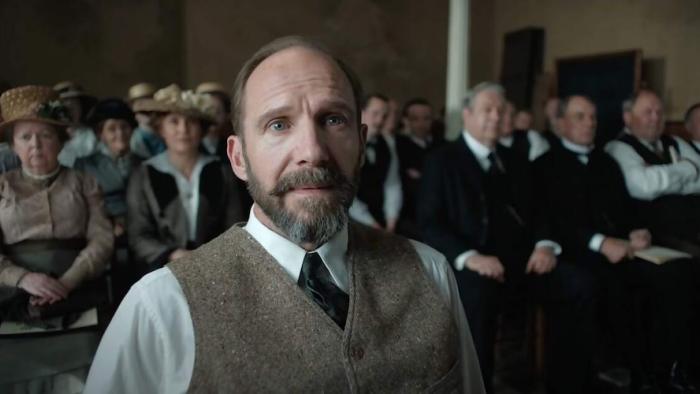 The Choral movie: A bearded Ralph Fiennes in vest and tie stands in crowded hall