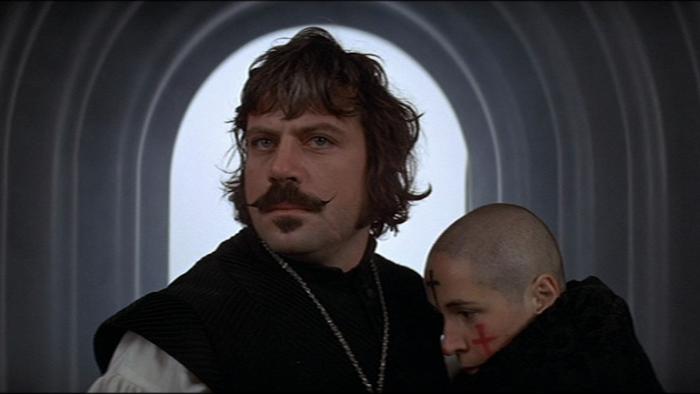 The Devils: Man with mustache holding a woman with a shaved head in a hallway