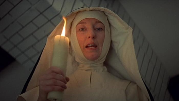 The Devils: Nun holding a lit candle, gazing forward, in a dimly-lit room