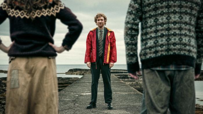 The Incomer: Man in red jacket stands between two people, who are seen from behind