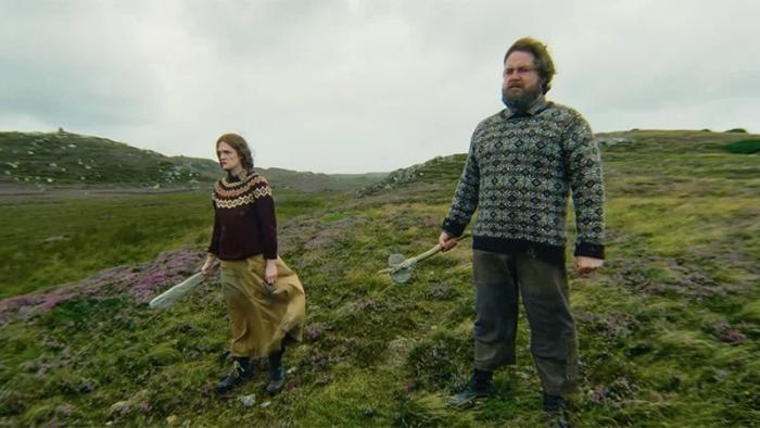 The Incomer: A woman and a man in sweaters stand on a grassy hill under a cloudy sky holding bats as weapons
