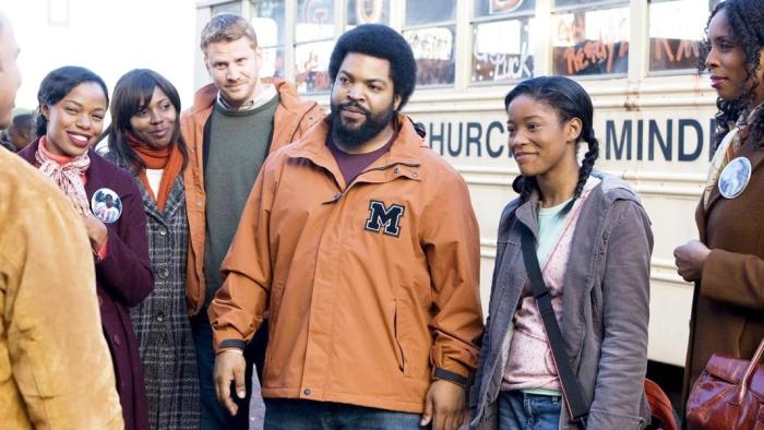 Ice Cube and Kiki Palmer stand in front of a school bus in casual clothing talking with a group of people.