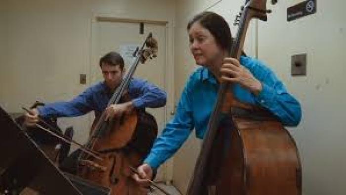 The Only Girl in the Orchestra: A woman and a man are shown from the waist up playing double basses together.