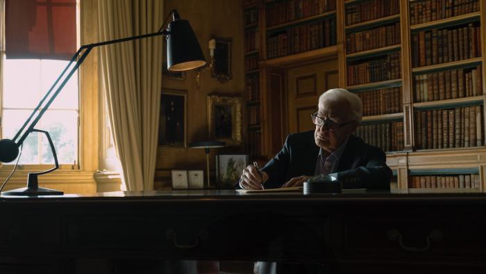 Still from The Pigeon Tunnel: John le Carré, the White British author who is the subject of the film, is seated at a desk with a black lamp while writing with a pen, the gray-haired writer is wearing a dark-colored sports jacket, a light-colored Oxford shirt with the collar opened, and gray pants, and in the background are bookshelves and framed images hanging on a wall or positioned on a nearby table