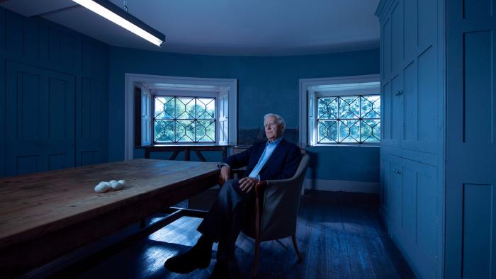 Still from The Pigeon Tunnel: John le Carré, the White British author who is the subject of the film, is seated in a chair with legs crossed and next to a long wooden rectangular-shaped table that has six white eggs on it, above is a fluorescent ceiling light, and the gray-haired writer is wearing a light-colored Oxford shirt with an open collar, and dark-colored sports jacket, pants, socks, and shoes, and in the background are two windows 
