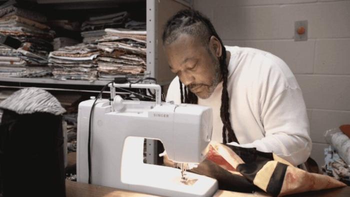 The Quilters: Chill, a man of color with long locks and grizzled beard, bends over a sewing machine preparing to seam some fabric.