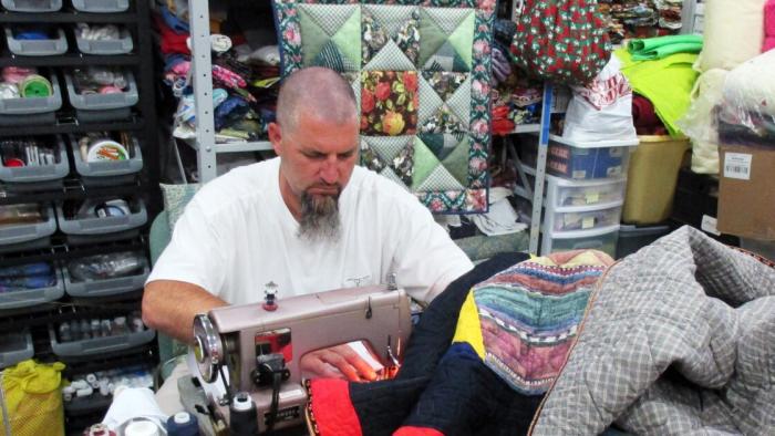 The Quilters: A White man with a shaved head and greying beard concentrates while using a sewing machine.