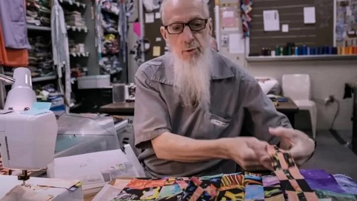 The Quilters: A White man with glasses and a long grey beard lays out strips of fabric preparing to piece a quilt together.