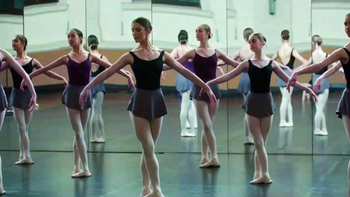 A line of young female ballet dancers pose identically in a mirrored dance studio