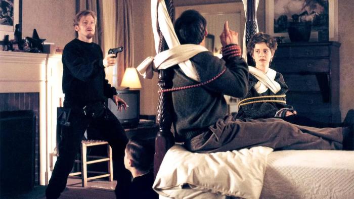 The Ref movie: In a bedroom, Denis Leary left points a gun at a man and woman tied up to a bed with a teenage boy sitting at the foot of the bed