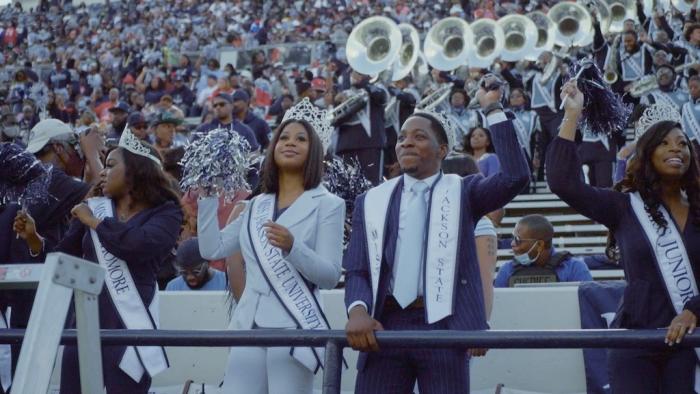 Still from The Return: In the foreground, three African American women standing in stadium bleachers, wearing business suits, royalty sashes, and crowns, and one Black man standing with the women, wearing business attire and a sash, at HBSC football game with other fans in the background