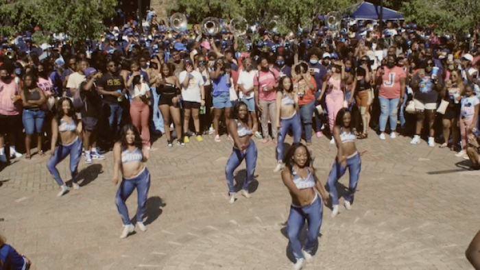 Still from The Return: Six African American women surrounded by a circle of onlookers perform dance steps, wearing light-colored halter tops, blue jeans, and white sneakers