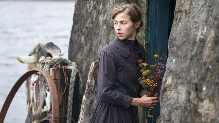 The Road Dance: A young woman stands looking over her shoulder, holing a bunch of flowers outside a cottage door