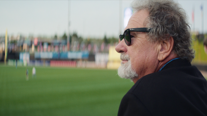 Still from The Saint of Second Chances: Left facial profile of Mike Veeck as an adult looking out a baseball stadium, wearing dark-colored sunglasses and dark jacket