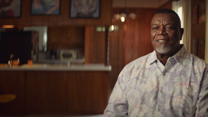 Still from The Saint of Second Chances: Firsthand interview with Lamar Johnson, a former Chicago White Sox player, faces and the camera and is seated, wearing a pastel floral oxford shirt unbuttoned at the top