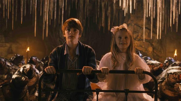 A brother and sister in their PJs sit atop their iron bed in an underground cavern
