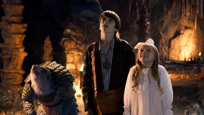 A brother and sister stand in an underground cavern next to a standing pangolin with a look of awe on their faces