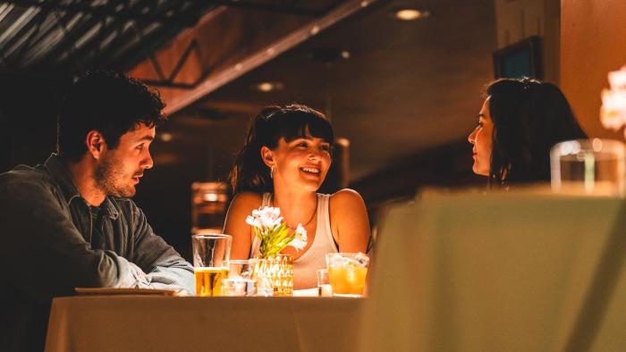 The Threesome movie: At a restaurant table, Josh Hauer-King looks at Zoey Deutch as she smiles across the table to Ruby Cruz