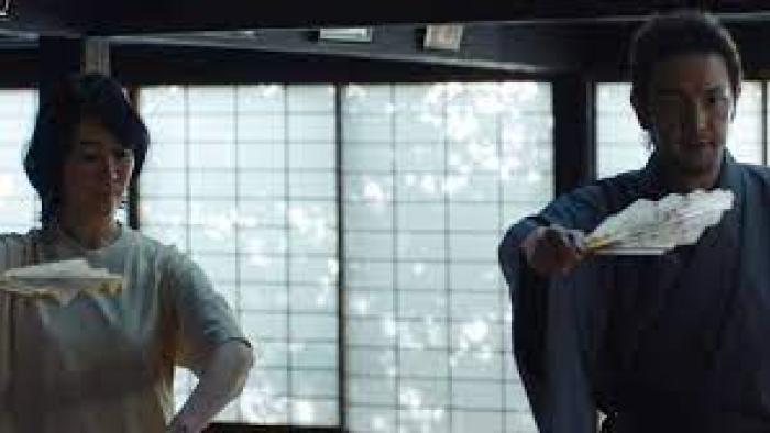 The Village (2023): A Japanese woman left and a Japanese man right each hold a fan horizontally in their left hands and face forwards as they practice a Japanese Noh dance performance