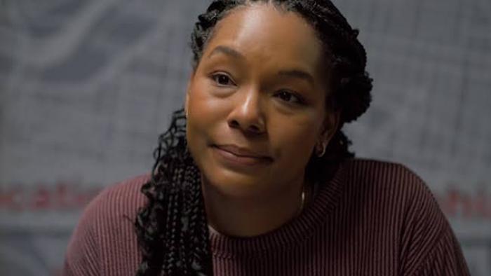 The Body in the Locker: Black Woman with braids sitting, wearing a maroon sweater, neutral expression.