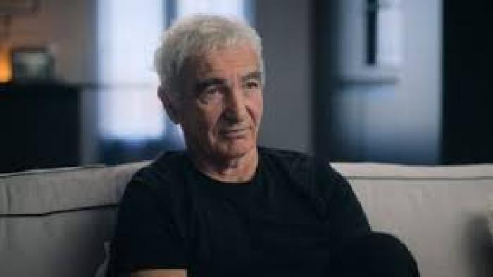 The Bus: A French Football Mutiny Movie: Older White man with grey hair sitting on a couch in a softly lit room, wearing a black shirt.
