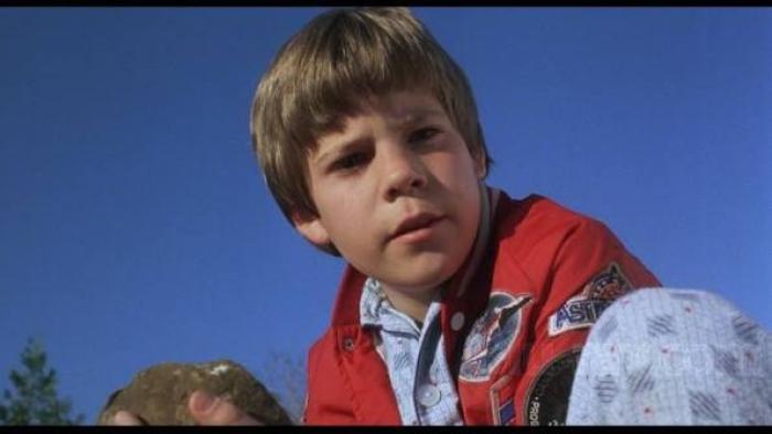 The Gate: Young boy in red jacket holding a rock, blue sky background.