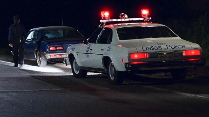 The Thin Blue Line: A police car is shown pulled over behind a blue car on the side of the road with a police officer walking toward the front car