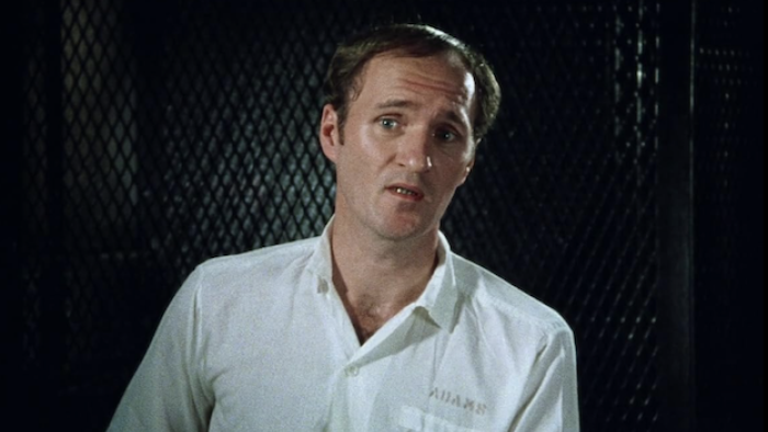 The Thin Blue Line: Man in white shirt stands by a dark textured background speaking to camera as part of an interview