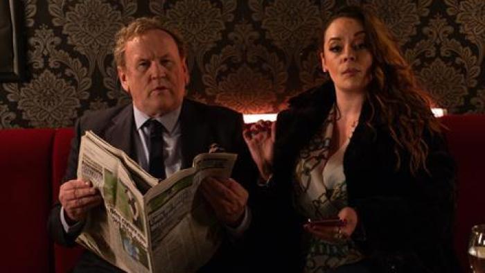 Three Day Millionaire: An older businessman in a suit holds a newspaper and his female partner looks at the camera