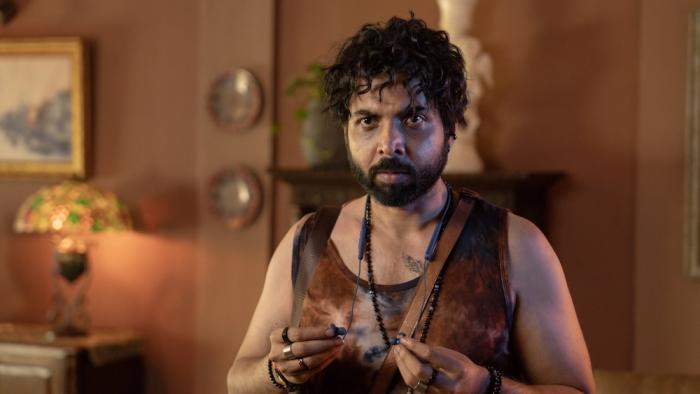 Toaster: An Indian man with curly hair and tank top holding some ear buds, standing in a warmly lit room