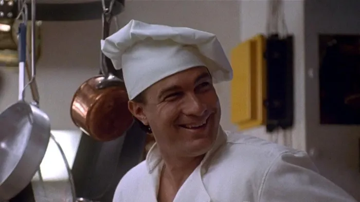 Under Siege: Chef in white uniform smiling in a kitchen with pots and pans hanging