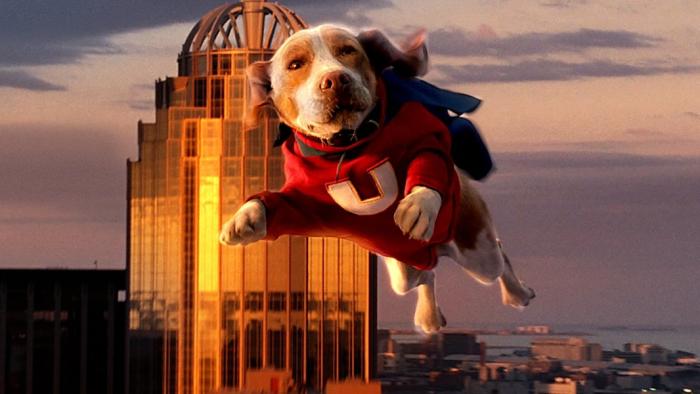 The Underdog, voiced by Jason Lee, leaps in the air wearing a red suit with a yellow U on the chest with a blue cape, a sunset cityscape in the background.