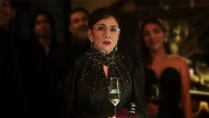 Uninvited movie: Filipino older woman in glasses and black sparkly dress holds champagne glass while at fancy party, other guests behind her