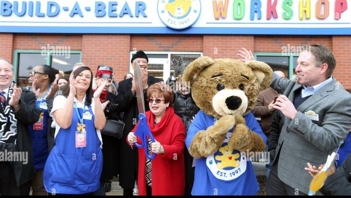 Unstuffed: A Build-a-Bear Story: A group of people and a costumed teddy bear at an outdoor ribbon-cutting ceremony.