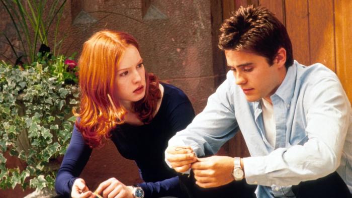 Urban Legend: Red-haired woman and brown-haired man sitting outdoors, engaged in conversation