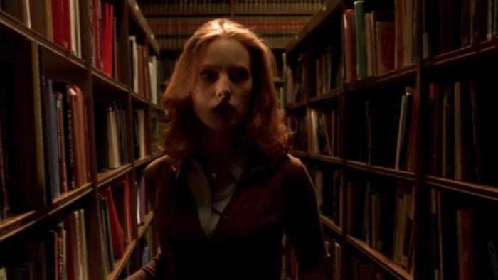 Urban Legend: White woman looks scared as she walks between bookshelves in a library in dim lighting
