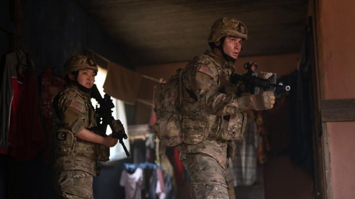 Valiant One movie: Chase Stokes stands in front of Lana Condor while aiming his gun
