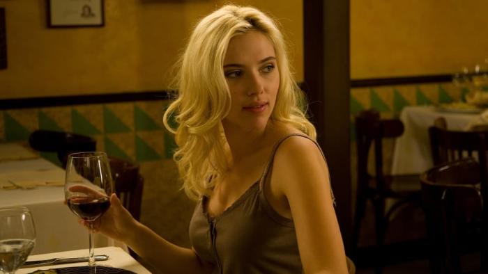 Scarlett Johansson sits at a restaurant table holding a glass of red wine, looking over her shoulder. 
