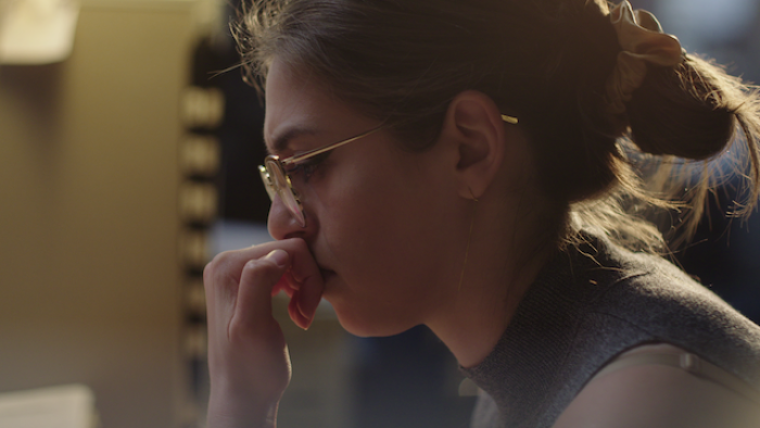 Still from Victim/Suspect: Profile of investigative reporter Rachel de Leon, a White journalist, seated at a newsroom cubicle and looking downward at 