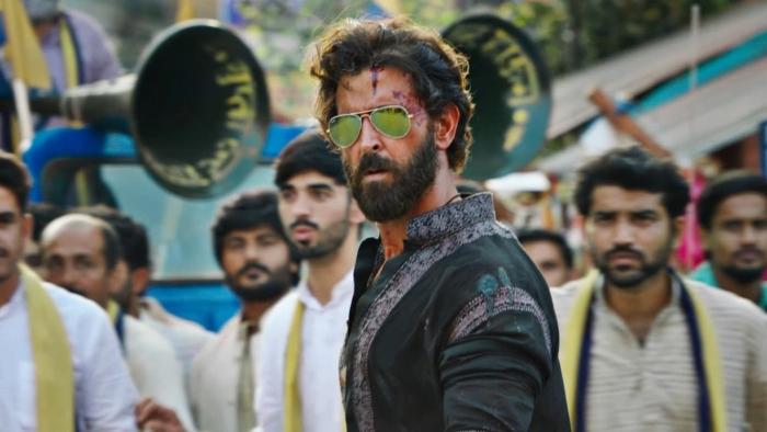 Vikram Vedha: A bearded Indian man wearing Aviator sunglasses looks on with a crowd of men behind him