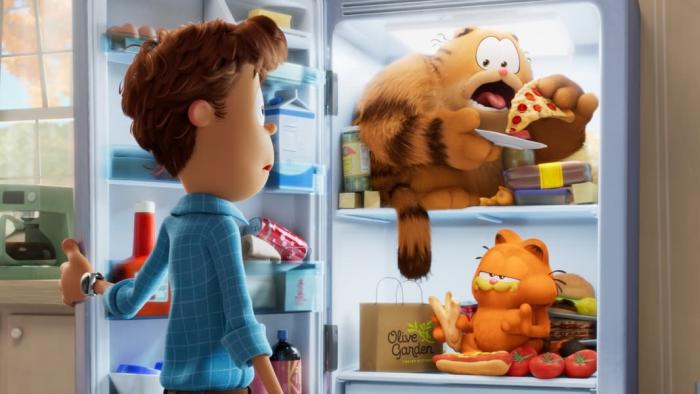The Garfield Movie: Jon stands in front of an open refrigerator, where Garfield and Vic sit on shelves eating the food