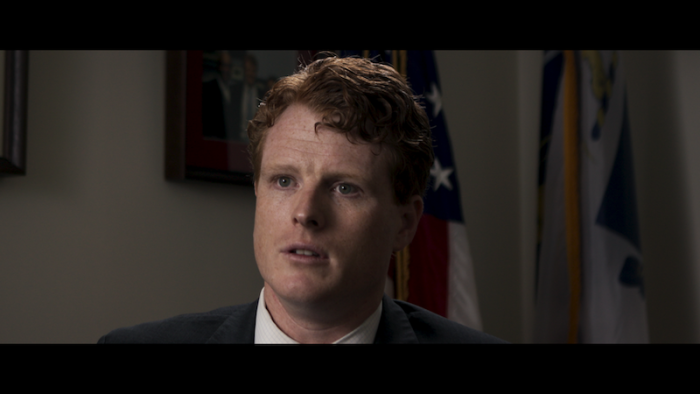 Still from Wake Up: Stories from the Frontlines of Suicide Prevention: Rep. Joe Kennedy III, a White U.S. Congressman, faces the camera and is seated for a firsthand interview, wearing a white shirt, dark-colored jacket, and reddish-brown colored hair