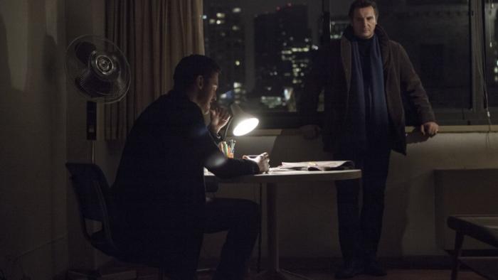 A Walk Among the Tombstones: Two men in dimly lit office, one seated at desk with lamp, other standing by a window.